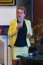 Kobieta w okularach w żółtej garsonce i czarnej bluzce przemawia stojąc za mównicą i trzymając mikrofon