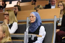 Uczestnicy siedzący w auli. Na pierwszym planie kobieta w okularach, białej koszuli, czarnej kamizelce i niebieskiej wzorzystej chuście na głowie, uważnie słuchająca.