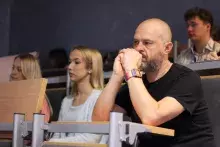 Mężczyzna z brodą i kolorowym zegarkiem siedzi w ławce w auli, opierając podbródek na splecionych dłoniach i uważnie słuchając. Obok niego widoczne są inne osoby.