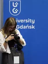 Kobieta w okularach trzymająca profesjonalny aparat fotograficzny i patrząca na jego ekran. Za nią znajduje się niebieski baner z logo i napisem "University Gdańsk".