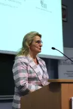prof. Urszula Patocka-Sigłowy w okularach i marynarce w kratę stoi za drewnianą mównicą, przemawiając do mikrofonu. Za nią znajduje się ekran projekcyjny.