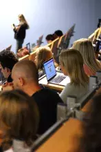 Widok zza pleców uczestników siedzących w auli. Wiele osób, niektóre z otwartymi laptopami, jest zwróconych w stronę przodu sali, gdzie w oddali stoi jedna osoba.