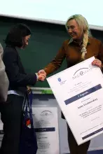 Uśmiechnięta kobieta w brązowej kurtce wręcza laureatce, Annie Grabowskiej, oprawiony dyplom za I miejsce w konkursie "medi@stery" i ściska jej dłoń.
