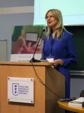 Kobieta w okularach i niebieskiej sukience stoi za drewnianą mównicą z logo Wydziału Nauk Społecznych UG i przemawia do mikrofonu.