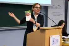Kobieta w okularach i apaszce stoi za mównicą, przemawia do mikrofonu i prezentuje publiczności książkę, trzymając ją w podniesionej ręce.
