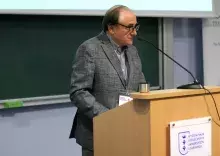 Mężczyzna w okularach i marynarce w kratę stoi za drewnianą mównicą z logo Wydziału Nauk Społecznych Uniwersytetu Gdańskiego i przemawia do mikrofonu.