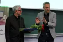 Profesor Harciarek w szarej marynarce przemawia i gestykuluje, stojąc obok profesora Treppy w okularach, który trzyma niebieską teczkę i bukiet czerwonych róż.