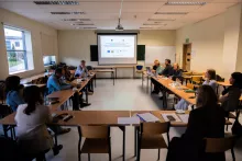 Szerokie ujęcie sali konferencyjnej, w której uczestnicy siedzą przy stołach ustawionych w kształcie litery U. Wszyscy patrzą w stronę ekranu projekcyjnego z przodu sali, na którym wyświetlana jest prezentacja.