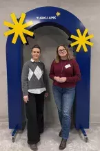 Dwie uśmiechnięte kobiety stoją obok siebie pod dużym, niebieskim łukiem dekoracyjnym z napisem „TURKU AMK” i żółtymi gwiazdami. Kobieta po lewej ma na sobie szary sweter w romby i czarne spodnie, a kobieta po prawej bordowy sweter, jeansy i plakietkę z imieniem „Dorota”.