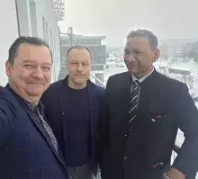 Zdjęcie typu selfie przedstawiające trzech mężczyzn (tych samych co na poprzednim zdjęciu) stojących na balkonie lub tarasie. Są ubrani w ciemne marynarki i płaszcze. W tle rozpościera się widok na zaśnieżony kampus uniwersytecki oraz budynki pod zachmurzonym, zimowym niebem