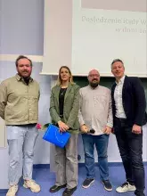 Zdjęcie przedstawiające 4 osoboy podczas wręczenia nagrody, 3 mężczyzn w strojach formalnych i kobieta w stroju formalnym trzymająca dyplom i czerwoną różę