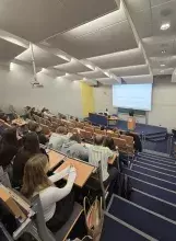 Widok z góry na wypełnioną studentami salę wykładową podczas prezentacji pt. „The army as tool of Rwandan policy (2003-2025)”. Prelegent stoi przy mównicy na dole sali, a słuchacze w rzędach przed nim robią notatki.