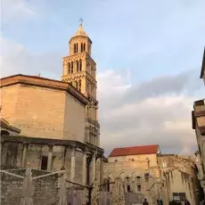 Widok zabytkowej uliczki z budynkami w kolorze piasku z czerwonymi dachami, widoczna wieża ze strzelistymi oknami