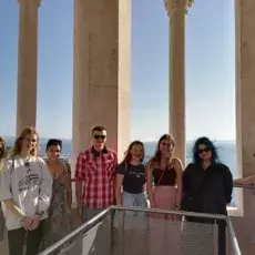 Grupa studentów na tle zabytkowych kolumn, na dalszym planie przymglony zielony pejzaż pod błękitnym niebem