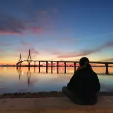 Ujęcie z tyłu studentki siedzącej na plaży, w tle morze o zachodzie słońca